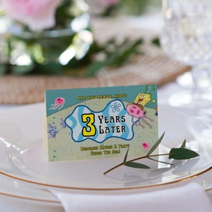 Peut inclure: Une carte de table avec une image de dessin anim&eacute; de Bob l'&eacute;ponge et une m&eacute;duse. Le texte sur la carte dit "&Ecirc;tes-vous pr&ecirc;ts, les enfants ? 3 ans plus tard. Benjamin organise une f&ecirc;te sous la mer !"