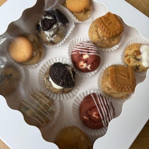 May include: A white box with a clear lid containing an assortment of gourmet cookies. The cookies include red velvet with white drizzle, cookies with Oreo pieces, and cookies topped with a Biscoff cookie. Each cookie is in a white paper cup.