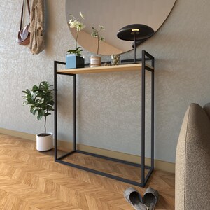 May include: A black metal console table with a wooden top shelf and a lower shelf. The table has a modern design and is perfect for a hallway or entryway. There is a small potted plant, a lamp, and a few decorative items on the top shelf.
