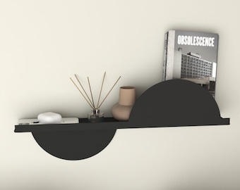 May include: Black metal shelf with two half-circle shelves. The shelf is mounted on a white wall and has a book titled "Obsolescence" and a small vase with dried sticks on it.