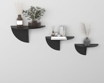 May include: Three black half-circle wall shelves with rounded edges. The top shelf has a glass bottle and a small potted plant. The middle shelf has a book, a candle, and a small vase. The bottom shelf has a vase with dried flowers.