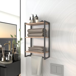 Puede incluir: Un estante de baño de metal negro con tres estantes de madera. El estante está montado en una pared blanca y tiene toallas y artículos de tocador.