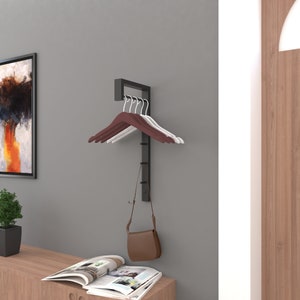 May include: A black metal wall-mounted coat rack with a shelf and hooks. The rack has a brown leather purse hanging from one of the hooks. There are also four wooden hangers on the rack, two brown and two white.