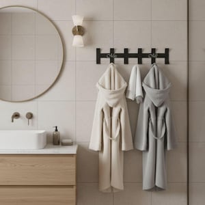 May include: A bathroom scene featuring a black wall-mounted coat rack with the words "Mrs." and "Mr." in script. Two hooded bathrobes, one cream and one gray, hang below. A round mirror and a modern sink are also visible.