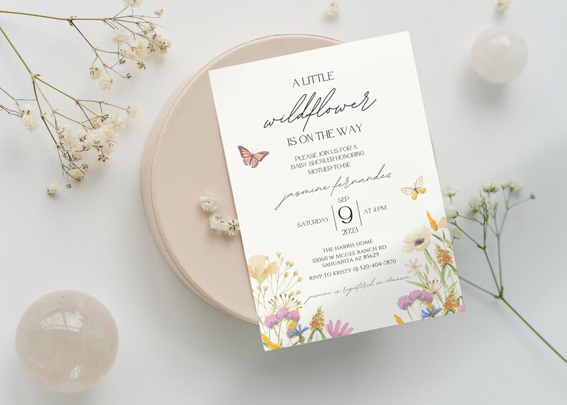 Puede incluir: Una invitaci&oacute;n de baby shower blanca con un borde floral y mariposas. El texto dice "A Little Wildflower is on the Way" e incluye el nombre "Jasmine Fernandes", la fecha "9 de septiembre de 2023" y la hora "4 PM".