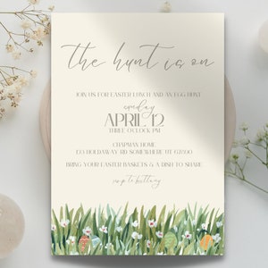 K&ouml;nnte beinhalten: Eine beige Einladungskarte mit dem Text "the hunt is on" in kursiver Schrift. Die Einladung ist f&uuml;r ein Osteressen und eine Ostereiersuche am Sonntag, den 12. April um 15:00 Uhr. Die Adresse lautet 123 Holdaway Rd Somewhere, UT 67000. Die Einladung bittet die G&auml;ste, ihre Osterk&ouml;rbe und ein Gericht zum Teilen mitzubringen. Der untere Teil der Karte zeigt eine Aquarell-Illustration von gr&uuml;nem Gras, wei&szlig;en Blumen und bunten Ostereiern.