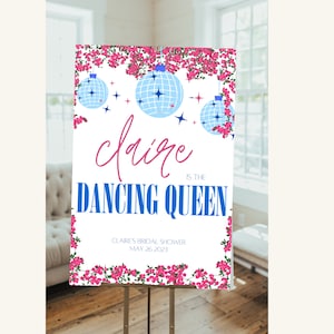 May include: A white sign with pink flowers around the border. The sign says "Claire is the Dancing Queen" with a blue disco ball graphic. The text "Claire's Bridal Shower May 26, 2023" is at the bottom of the sign.