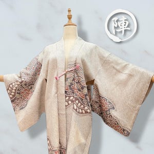 以下が含まれることがあります： 木製のマネキンに展示された、柄入りの袖とトリムが付いたベージュの着物。袖には茶色と赤の花柄のデザインがあります。着物はピンク色の留め具で留められています。日本語のテキストが入った白い円が右上隅にあります。