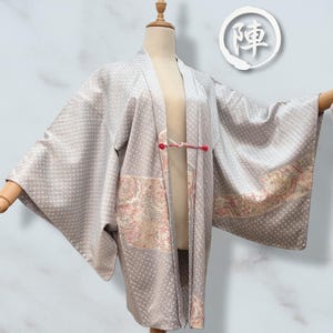 以下が含まれることがあります： 水玉模様と装飾的な花柄パネルが特徴の、明るい灰色の着物。着物はマネキンに展示されており、赤い留め具が付いています。日本語の文字が入った白い円形のエンブレムが見えます。