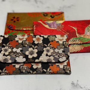 以下が含まれることがあります： 花柄と鳥柄の装飾的な布製の小さなポーチが3つ。ポーチは金、赤、黒の色合いで、複雑な模様が施されています。小物を入れるのに使われる可能性が高いです。