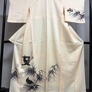 May include: Cream-colored kimono with a detailed illustration of birds and bamboo. The artwork is rendered in black and gray ink, creating a striking contrast against the soft background. The kimono is displayed on a black rack.