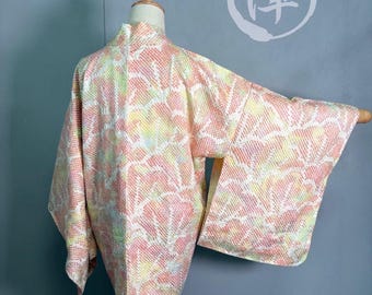 Puede incluir: Un kimono de color pastel con un estampado floral se exhibe en un maniquí. El kimono presenta una mezcla de tonos rosas, amarillos y blancos. Las mangas son anchas y la tela parece texturizada. Un carácter japonés es visible en el fondo.