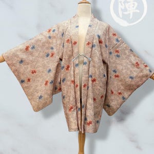 以下が含まれることがあります： 赤と青の花柄のベージュ色の着物。着物は木製のマネキンに展示されています。日本語の文字が右上隅にあります。着物は広い袖とゆったりとしたフィット感があります。