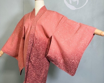 Puede incluir: Un kimono de color coral con un efecto degradado, que transiciona de un tono más claro en la parte superior a un borgoña moteado más profundo en la parte inferior. La prenda se exhibe en un maniquí de madera, mostrando su diseño abierto y mangas anchas.