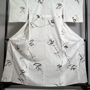 以下が含まれることがあります： 白地に縦縞模様と抽象的な黒とグレーの筆使いのデザインが施された着物。 着物は黒い金属製のラックに展示されており、その全長と流れるような生地が際立っています。