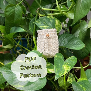 May include: A crocheted drawstring bag in a cream color with a wooden bead accent. The bag is hanging from a green leafy plant. The text "PDF Crochet Pattern" is written in a white cloud shape.