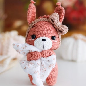 Może przedstawiać: Pluszowa zabawka w kształcie różowego króliczka z białą buzią i brązową kokardką. Króliczek trzyma w łapkach biały i różowy kocyk.