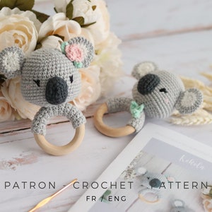 Peut inclure: Deux hochets en forme de koala en crochet gris avec des anneaux en bois. Le koala à gauche a une fleur rose sur la tête. Le koala à droite a une feuille verte sur la tête. Les jouets sont sur une surface en bois blanc avec un bouquet de fleurs blanches et une pelote de laine grise.