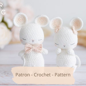 May include: Two white crocheted mouse amigurumi toys with pink ears and bows. The mouse on the left is wearing a bow tie, and the mouse on the right is wearing a bow. The toys are sitting on a white wooden surface. Patron - Crochet - Pattern