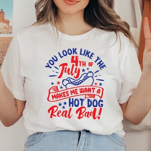 May include: White t-shirt with a graphic design in red and blue that reads "YOU LOOK LIKE THE 4TH OF JULY MAKES ME WANT A HOT DOG Real Bad!". The shirt is worn by a person.