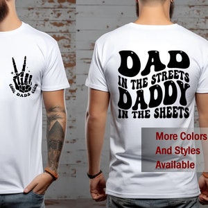 May include: White t-shirt with a black graphic of a skeleton hand making a peace sign with the text "Cool Dads Club". The back of the shirt has black text that reads "Dad in the Streets Daddy in the Sheets".