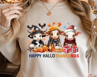 Könnte beinhalten: Ein weißer Pullover mit einem Aufdruck von drei Cartoon-Kühen, die in Kostümen für Halloween, Thanksgiving und Weihnachten verkleidet sind. Der Text "Happy HalloThanksmas" ist unter den Kühen aufgedruckt.