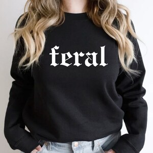 Puede incluir: Sudadera negra con la palabra "feral" impresa en blanco en una fuente g&oacute;tica.