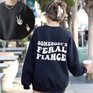 May include: Black sweatshirt with white text that reads "Somebody's Feral Fiance" on the back. The front of the sweatshirt has a white peace sign with a skeleton hand.