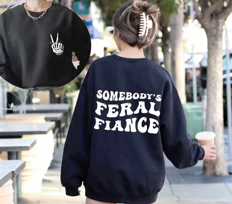 May include: Black sweatshirt with white text that reads "Somebody's Feral Fiance" on the back. The front of the sweatshirt has a white peace sign with a skeleton hand.