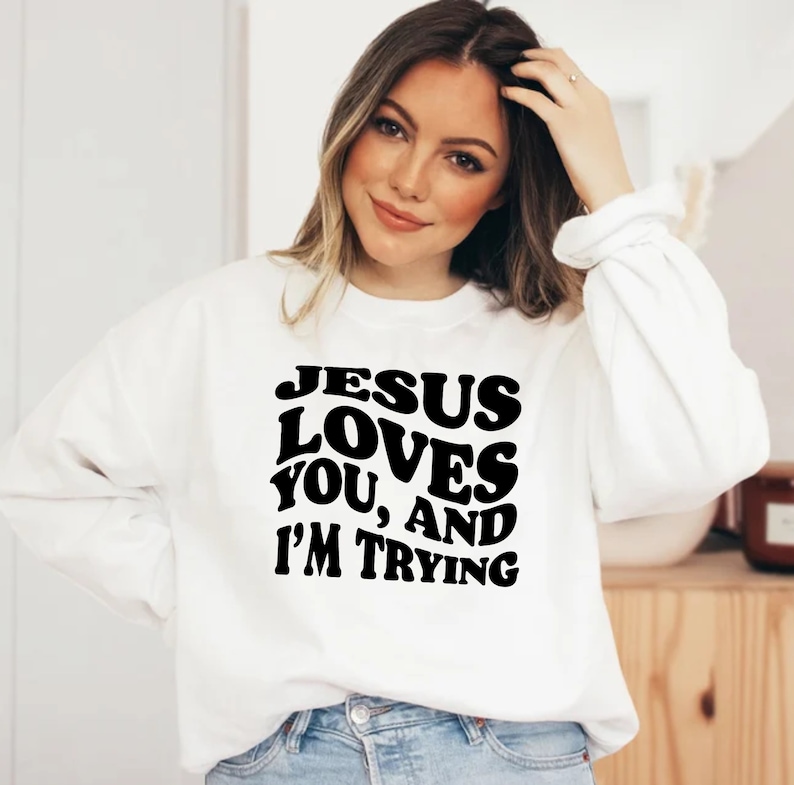 Pu&ograve; includere: Una felpa bianca con la scritta "JESUS LOVES YOU, AND I'M TRYING" in grassetto, nero, con un font retr&ograve;. La felpa ha maniche lunghe e girocollo.