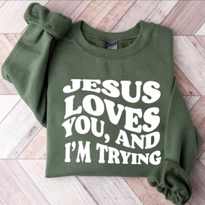 Pu&ograve; includere: Una felpa verde oliva con la scritta bianca "JESUS LOVES YOU, AND I'M TRYING" in un font retr&ograve;. La felpa ha un girocollo e maniche lunghe.