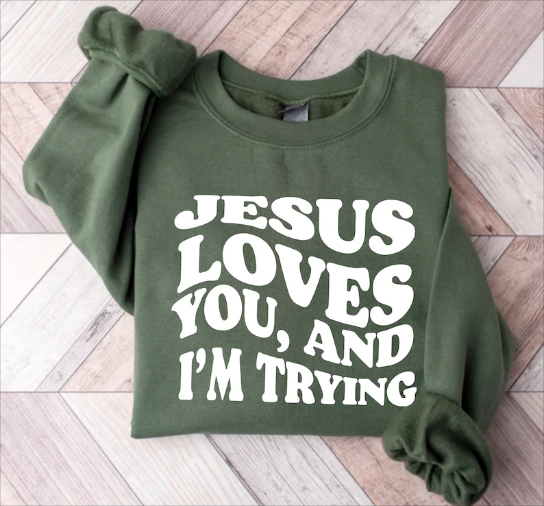Pu&ograve; includere: Una felpa verde oliva con la scritta bianca "JESUS LOVES YOU, AND I'M TRYING" in un font retr&ograve;. La felpa ha un girocollo e maniche lunghe.
