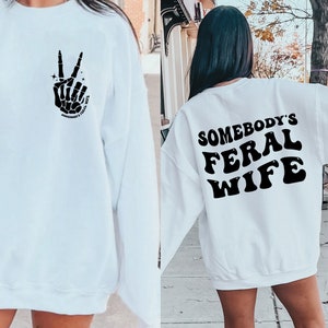 May include: White sweatshirt with a black skeleton hand making a peace sign on the front and the text "Somebody's Feral Wife" on the back.