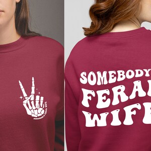 May include: A maroon sweatshirt with a white skeleton hand making a peace sign on the front and the text "Somebody's Feral Wife" in white on the back.