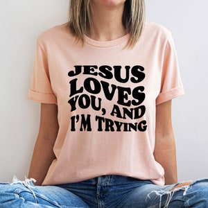 Pu&ograve; includere: T-shirt color pesca chiaro con maniche arrotolate. La maglietta presenta la scritta "JESUS LOVES YOU, AND I'M TRYING" in grassetto nero, con un lettering in stile retr&ograve;. La persona indossa jeans blu.