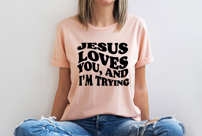 Pu&ograve; includere: T-shirt color pesca chiaro con maniche arrotolate. La maglietta presenta la scritta "JESUS LOVES YOU, AND I'M TRYING" in grassetto nero, con un lettering in stile retr&ograve;. La persona indossa jeans blu.