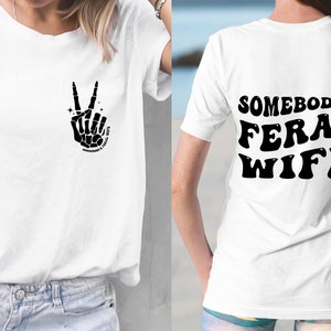 May include: White t-shirt with a black skeleton hand making a peace sign on the front and the text "Somebody's Feral Wife" in black on the back.