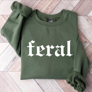 Puede incluir: Una sudadera verde con la palabra "feral" impresa en blanco en una fuente g&oacute;tica.