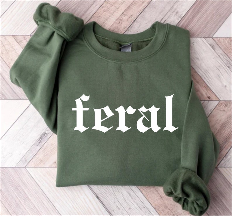 Puede incluir: Una sudadera verde con la palabra "feral" impresa en blanco en una fuente g&oacute;tica.