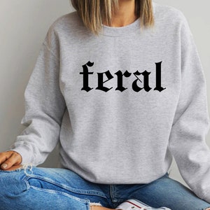 Puede incluir: Una sudadera gris con la palabra "feral" impresa en letras g&oacute;ticas negras.