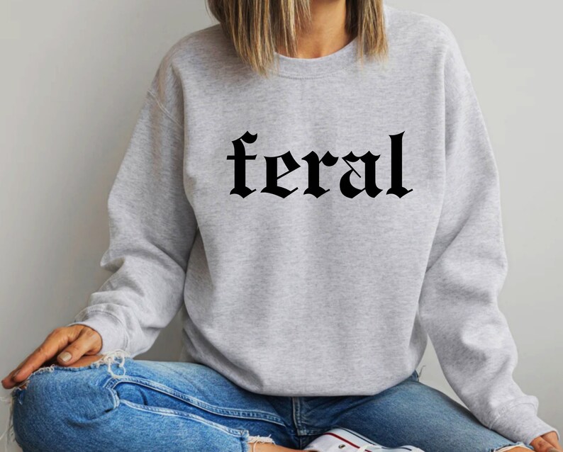 Puede incluir: Una sudadera gris con la palabra "feral" impresa en letras g&oacute;ticas negras.