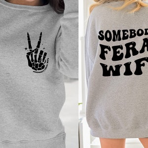 May include: Gray sweatshirt with a skeleton hand making a peace sign and the text "Somebody's Feral Wife" on the front. The back of the sweatshirt has the text "Somebody's Feral Wife" in a retro font.