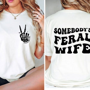 May include: White t-shirt with a black skeleton hand making a peace sign on the front and the text "Somebody's Feral Wife" in black on the back.