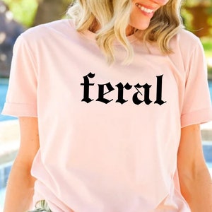 Puede incluir: Una camiseta rosa con la palabra "feral" impresa en letras g&oacute;ticas negras.