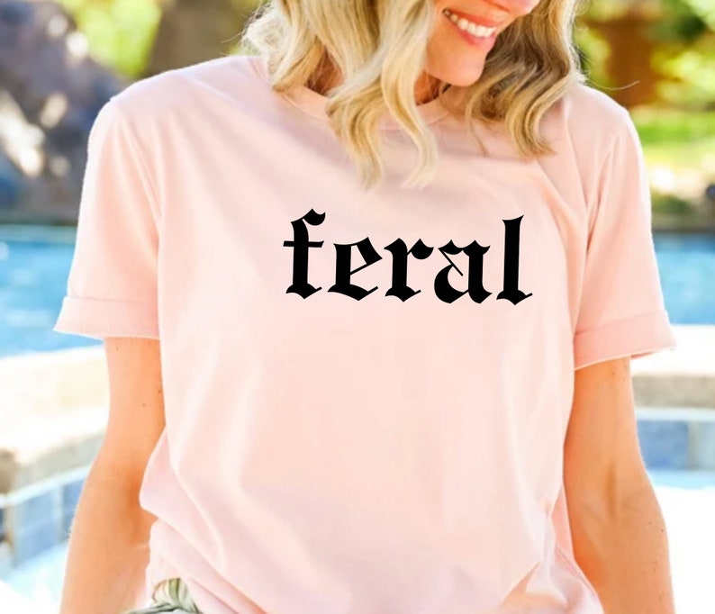 Puede incluir: Una camiseta rosa con la palabra "feral" impresa en letras g&oacute;ticas negras.