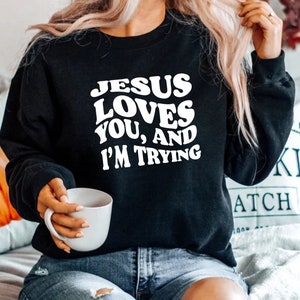Pu&ograve; includere: Felpa nera con la scritta bianca "JESUS LOVES YOU, AND I'M TRYING". Il testo &egrave; in grassetto e arrotondato. Una persona tiene in mano una tazza bianca. La felpa &egrave; un capo casual.