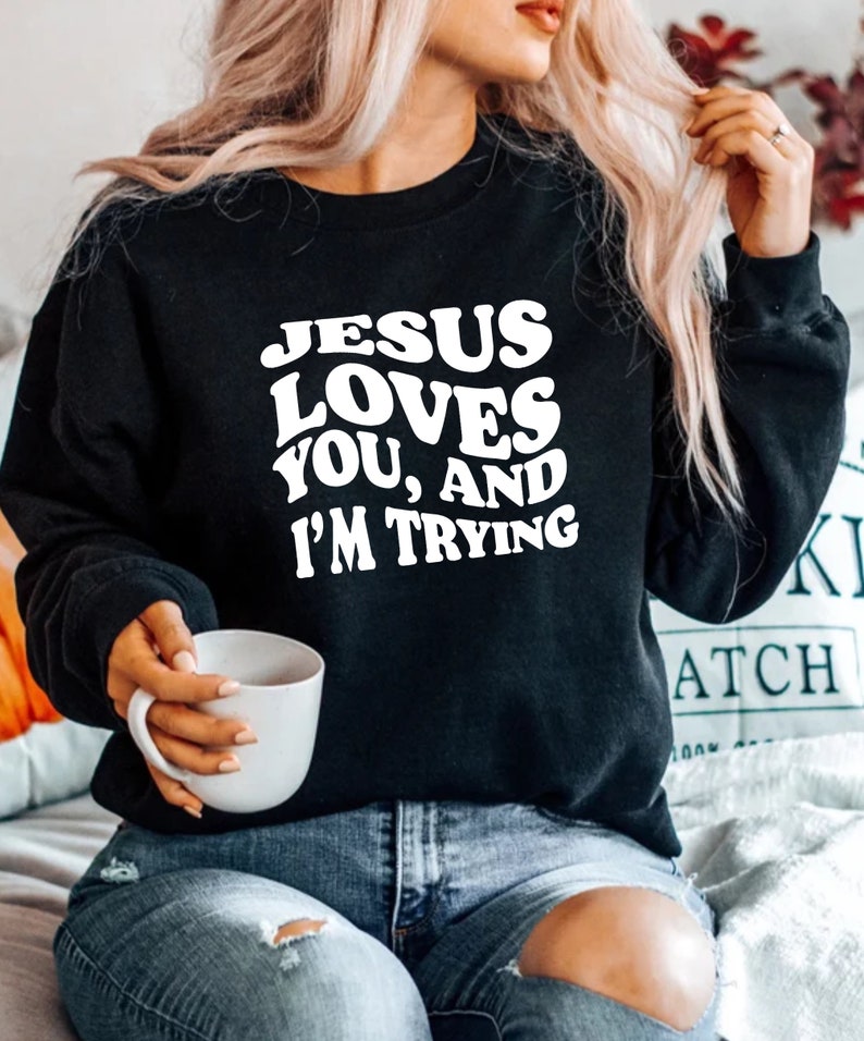 Pu&ograve; includere: Felpa nera con la scritta bianca "JESUS LOVES YOU, AND I'M TRYING". Il testo &egrave; in grassetto e arrotondato. Una persona tiene in mano una tazza bianca. La felpa &egrave; un capo casual.