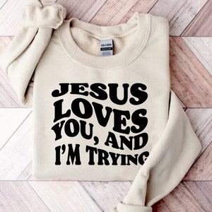 Pu&ograve; includere: Una felpa beige con la scritta "JESUS LOVES YOU, AND I'M TRYING" in grassetto nero. La felpa &egrave; piegata ordinatamente su una superficie di legno chiaro. Le maniche sono piegate e riposte.