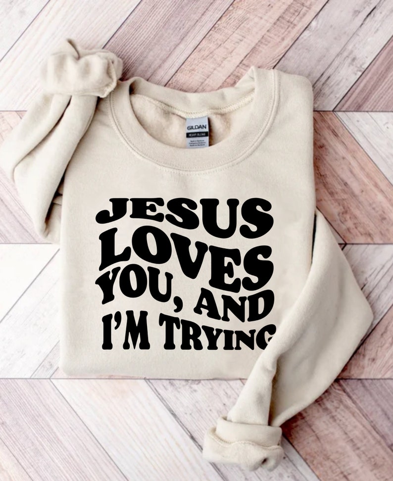 Pu&ograve; includere: Una felpa beige con la scritta "JESUS LOVES YOU, AND I'M TRYING" in grassetto nero. La felpa &egrave; piegata ordinatamente su una superficie di legno chiaro. Le maniche sono piegate e riposte.