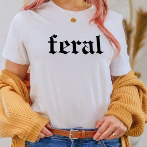 Puede incluir: Una camiseta blanca con la palabra "feral" impresa en letras g&oacute;ticas negras. La camiseta la lleva puesta una persona con el pelo rosa y un c&aacute;rdigan amarillo.